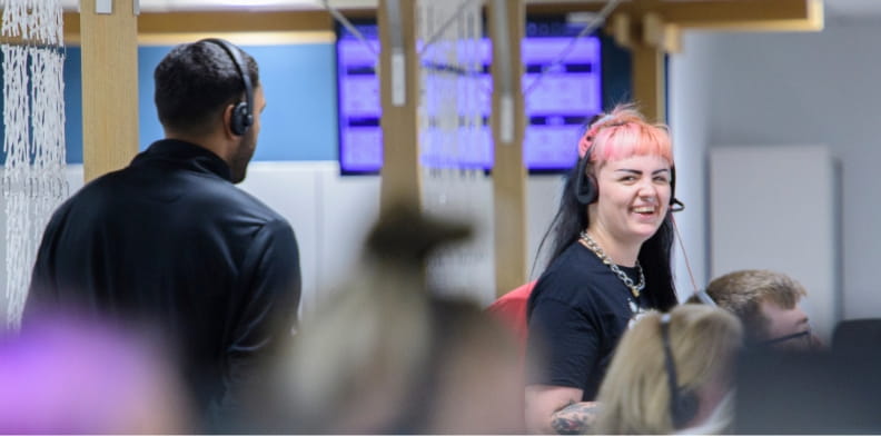 Working at HomeLet. This image displays a group of people out of focus working in the foreground with someone with pink hair in the background smiling directly to camera.