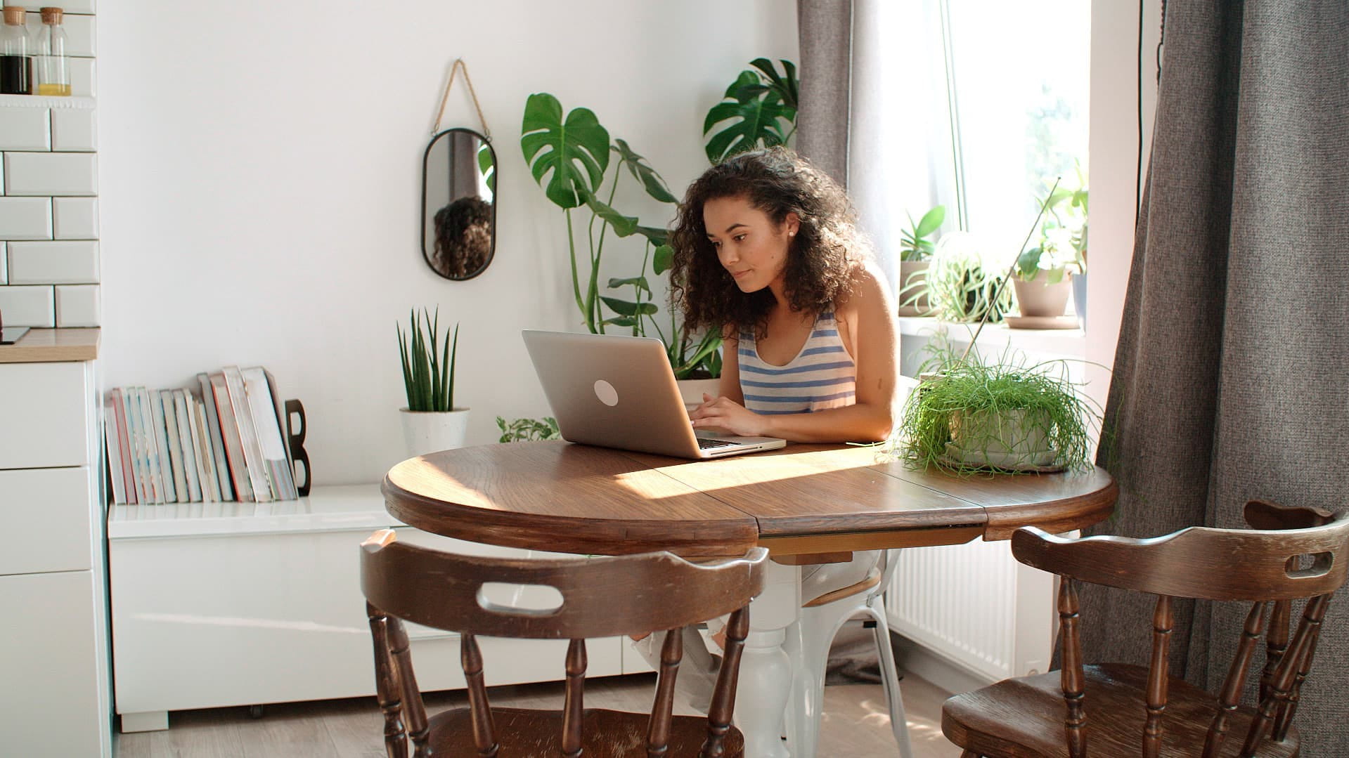 This image shows a woman sitting at a circular table at home, working on a laptop. There is a sunlit window behind her and plants around the room.