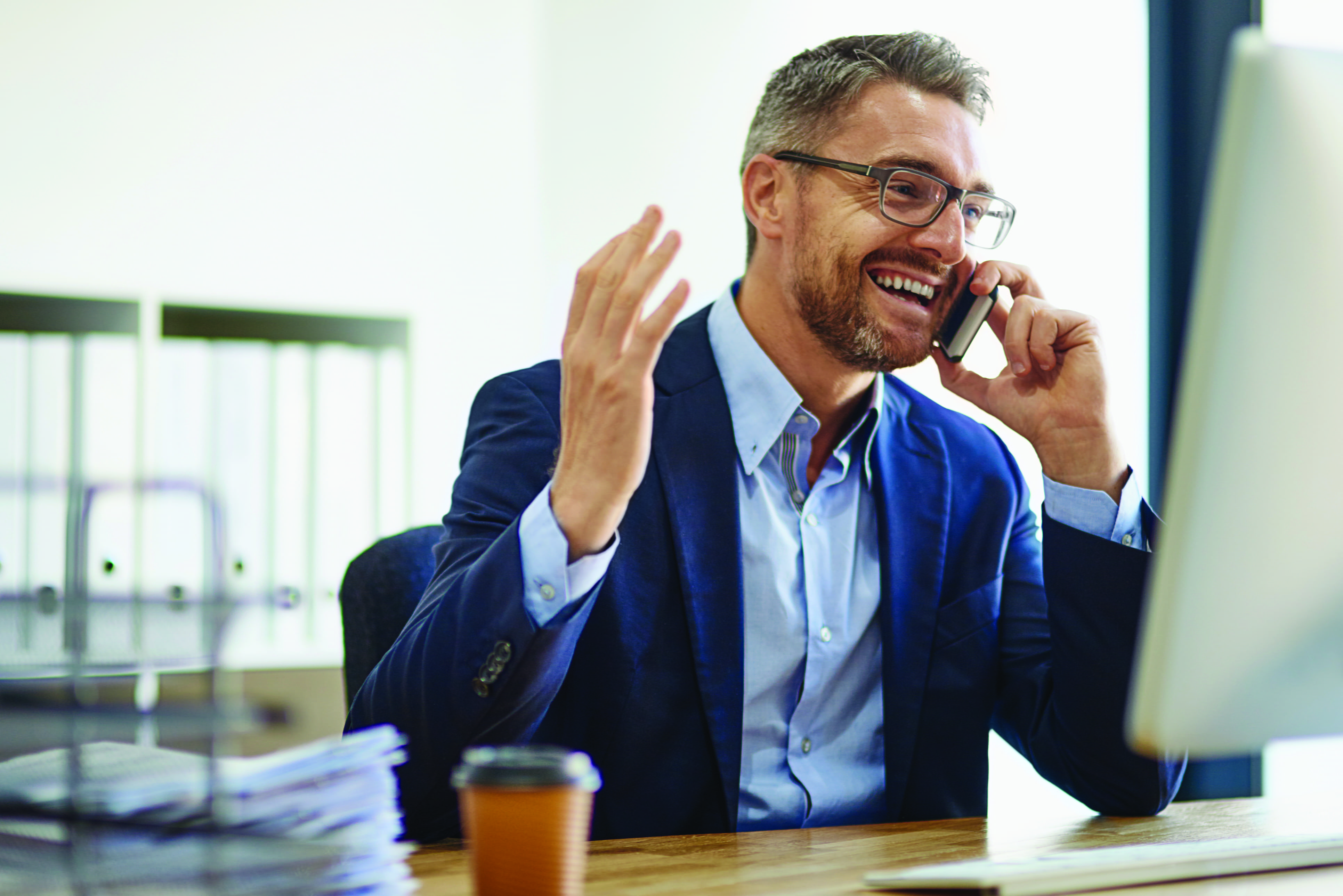 A person smiling and talking on their phone. In front of them is a table or desk with a pile of paper, a coffee cup and a monitor.