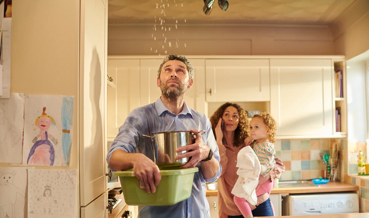 This photograph shows a family of 3. The dad is in the foreground catching water with a pan and washing up bowl while the mother looks on holding a small child.