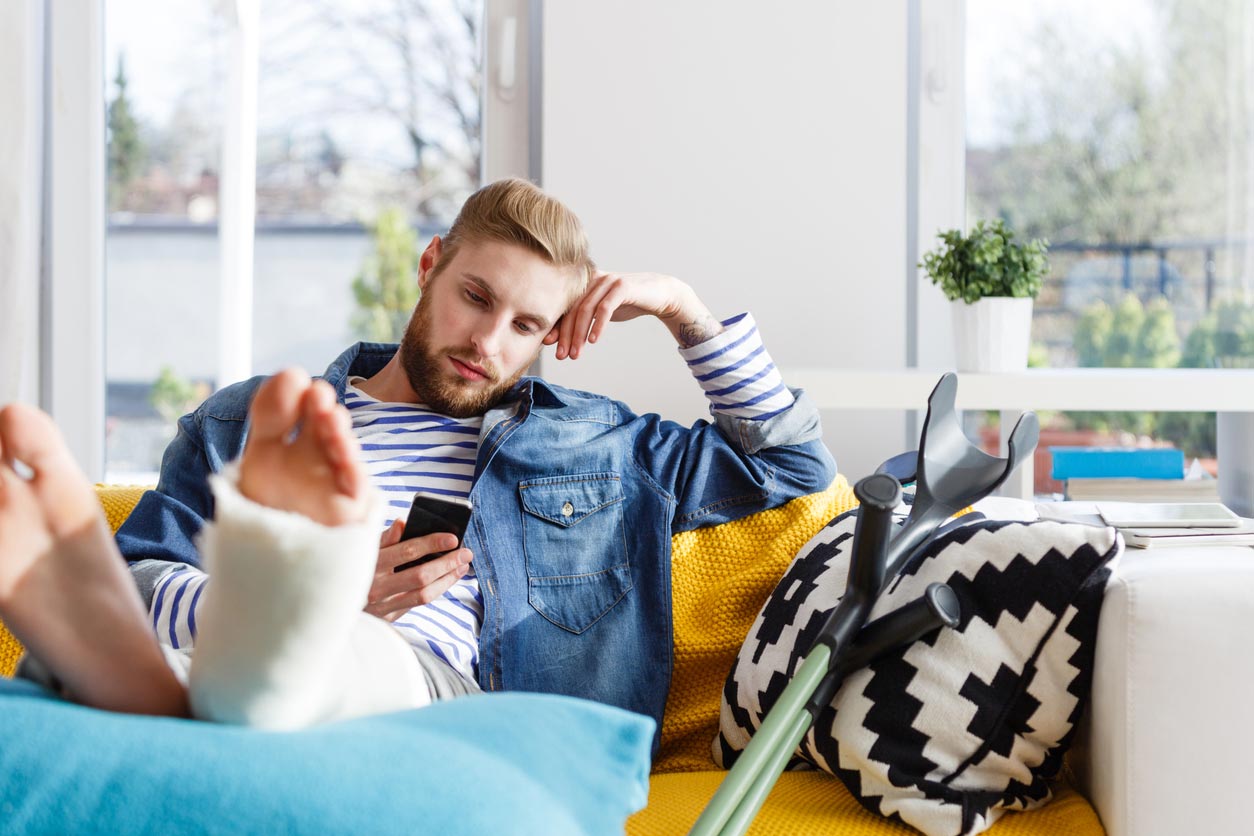 This photo shows a man sitting on a yellow sofa propped up by cushions, you can see crutches next to him and one of his feet is wrapped in plaster.