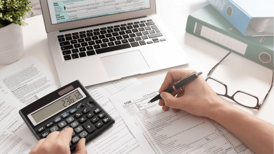 A photograph of a person writing on a form whilst also typing into a calculator. There is an open laptop in front of them and you can only see their hands.