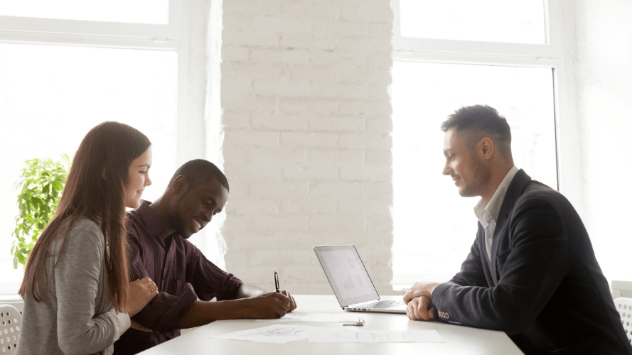 Tenants signing a tenancy agreement, while their landlord smiles at them from across a table.