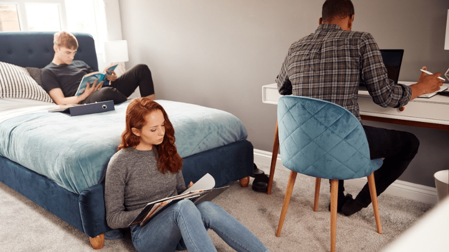 This photo shows three people, one is lying on a bed, another is sitting on the floor leaning against the bed and the third is working at a desk next to the bed.