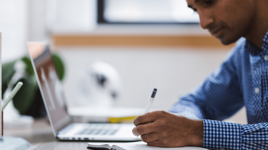 A photo of someone writing on a pad of paper with a laptop open next to them.