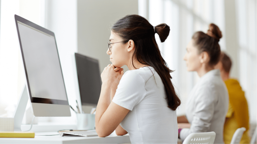 A rear shot of a person looking at a computer monitor, there are 2 more people in the background of the photo.